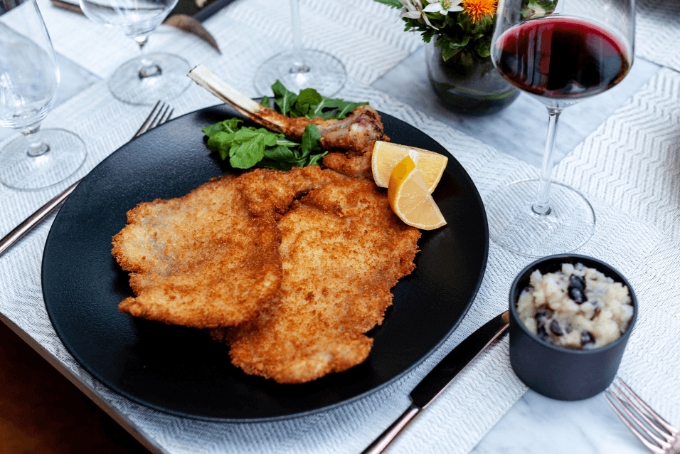 Breaded veal cutlets with lemon wedges on a black plate beside a glass of red wine