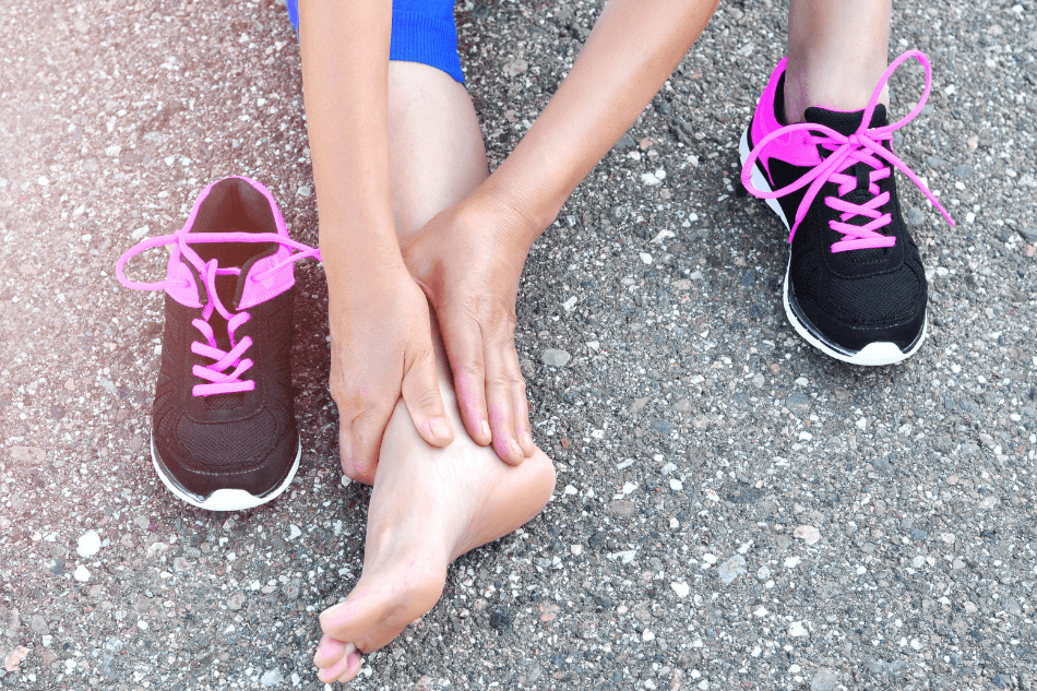 Runner sitting on pavement holding a bare foot between two black and pink running shoes